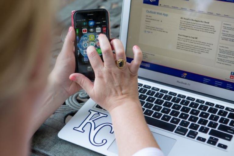 Woman logging into KU Email on a laptop and holding a smart phone that is showing a home screen with a list of applications